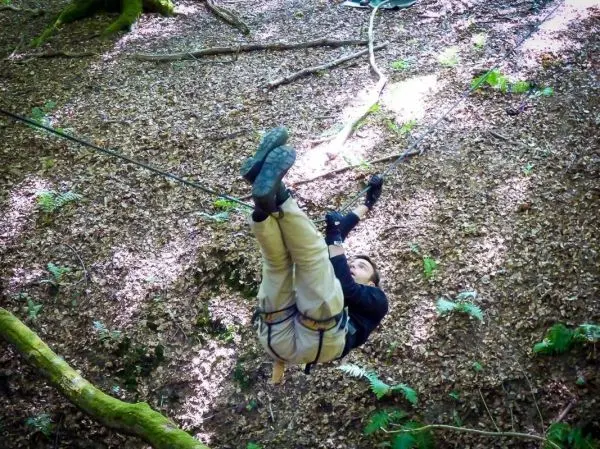 Seilquerung mit Sicherheitsgurt im Wald, Beine angezogen, Hände am Seil
