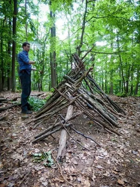 Selbstgebaute Laubhütte aus Ästen im Wald, Person steht daneben und hält Zweige