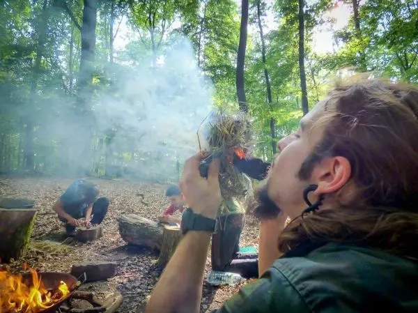 Zunder glimmt in der Hand, Rauch steigt auf, Feuer brennt im Hintergrund