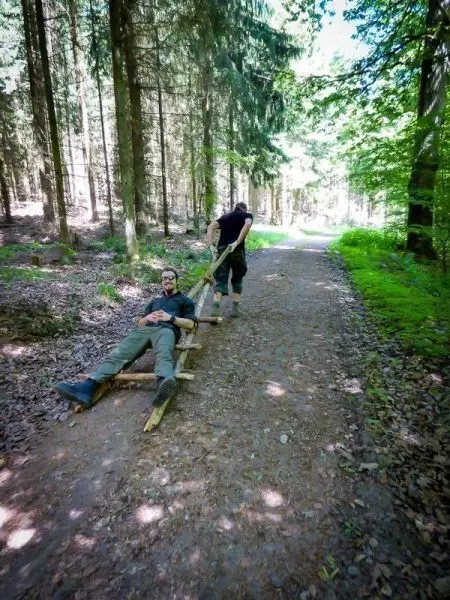 Zwei Personen transportieren eine improvisierte Trage durch den Wald