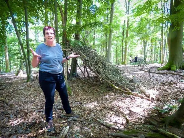 Frau steht in einem Wald neben einer selbstgebauten Notunterkunft aus Ästen und Laub