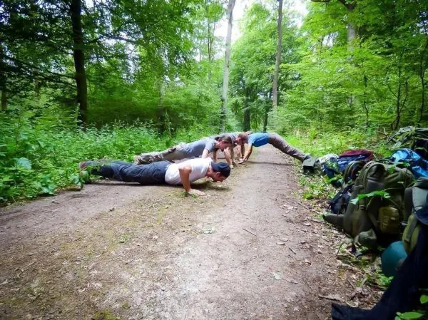 Gruppierung von vier Personen macht Liegestütze auf einem Waldweg, Rucksäcke im Hintergrund