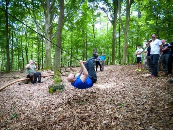 Mann überquert selbstgebaute Seilbrücke im Wald, Zuschauer beobachten