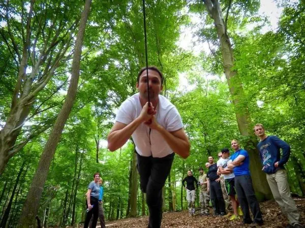 Mann überquert selbstgebaute Seilbrücke zwischen Bäumen, Zuschauer beobachten