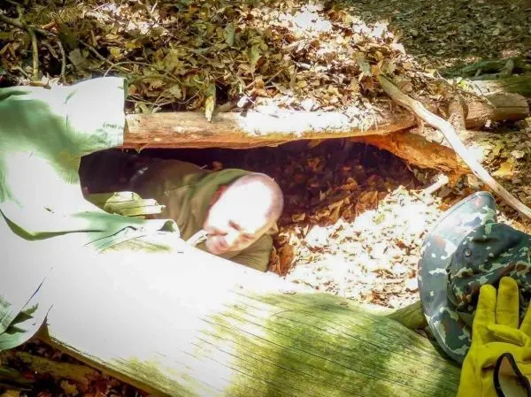 Person kriecht aus einem Erdloch unter Ästen und Laub