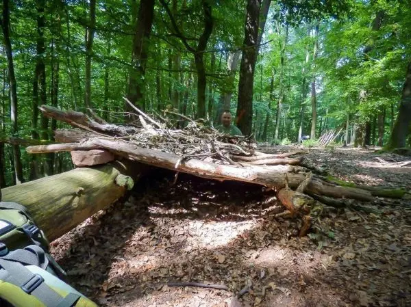 Person sitzt hinter einer selbstgebauten Unterkunft aus Ästen im Wald