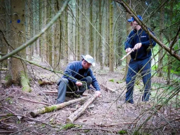 Zwei Personen bearbeiten Holzstücke im Wald, einer schnitzt mit einem Messer