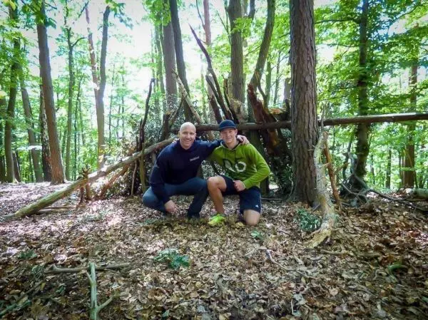 Zwei Personen posieren vor einer selbstgebauten Tarp-Notunterkunft im Wald
