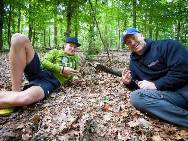 Zwei Personen sitzen im Wald auf dem Boden und zeigen mit den Händen