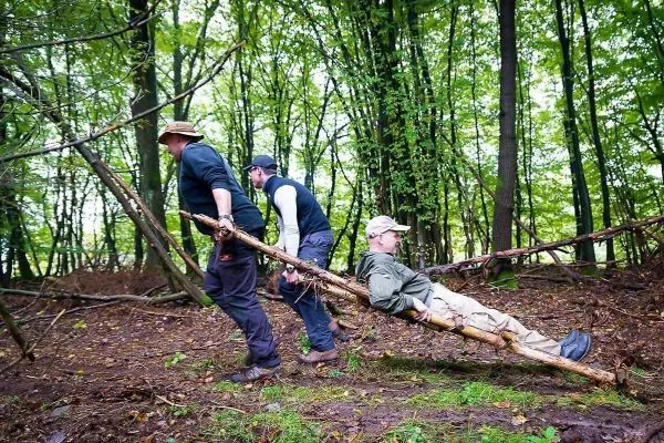Drei Personen ziehen eine improvisierte Trage aus Ästen durch den Wald