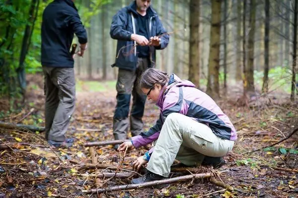 Frau sortiert Äste auf dem Boden im Wald, während zwei Personen im Hintergrund arbeiten