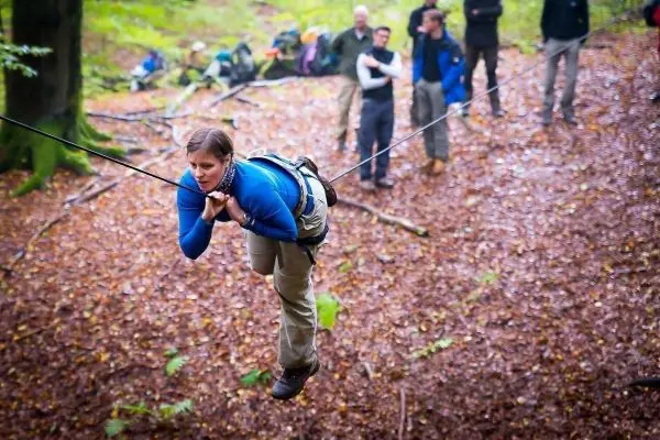 Frau überquert selbstgebaute Seilbrücke im Wald, Zuschauer beobachten