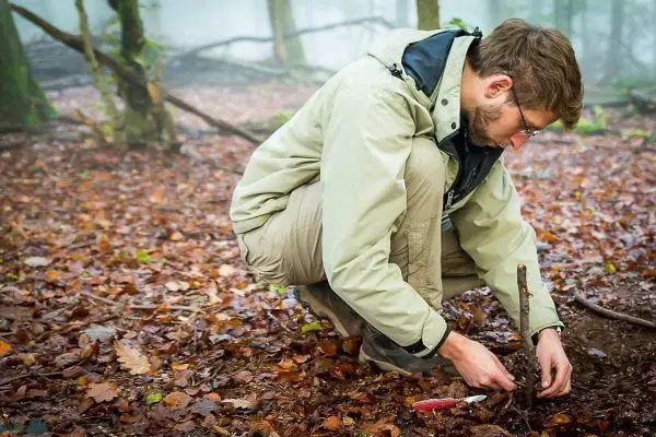 Mann kniet im Wald und bearbeitet einen Stock mit einem Messer