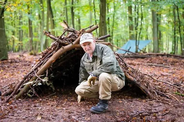 Mann kniet vor einer selbstgebauten Laubhütte im Wald