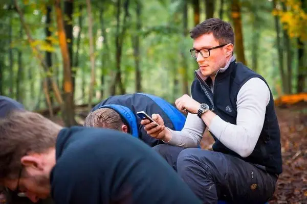 Mann sitzt im Wald und nutzt ein Smartphone, während andere sich bücken