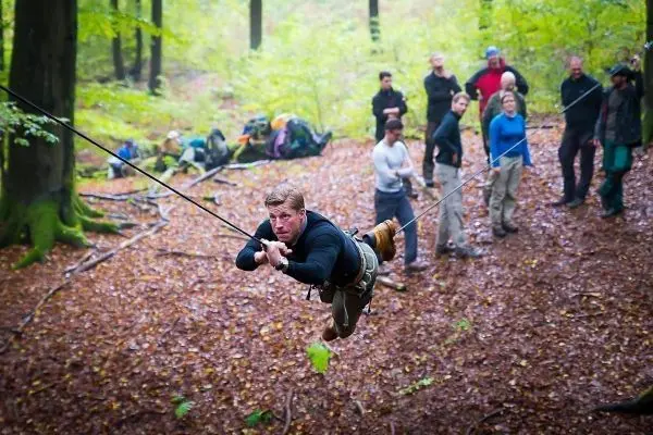 Mann überquert selbstgebaute Seilbrücke im Wald, Zuschauer beobachten