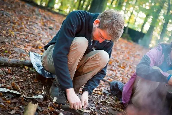 Person sitzt auf dem Boden und bearbeitet Holzstücke beim Überlebenstraining