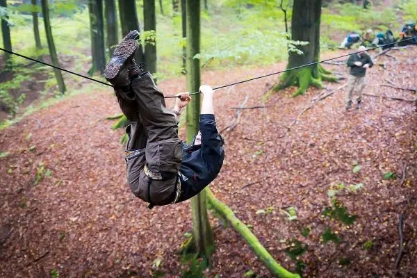 Person überquert selbstgebaute Seilbrücke im Wald, während andere im Hintergrund stehen