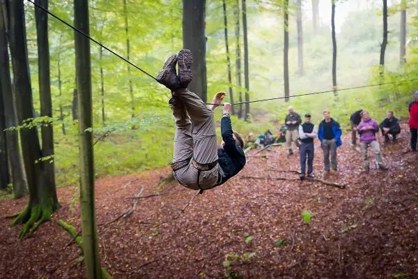 Person überquert selbstgebaute Seilbrücke im Wald, Zuschauer beobachten