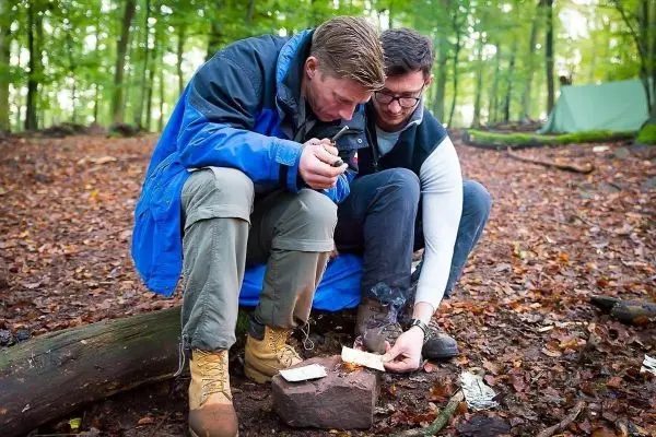 Zunder glimmt auf einem Stein, während zwei Personen konzentriert darüber gebeugt sind