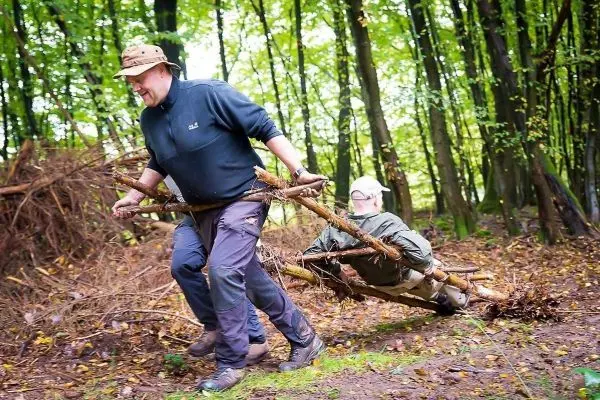 Zwei Personen transportieren eine improvisierte Trage aus Ästen durch den Wald