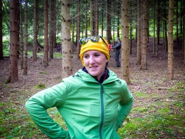 Frau in grüner Jacke und gelber Mütze steht im Wald zwischen Bäumen