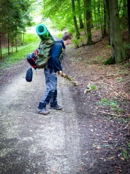 Junge hebt einen Stock auf einem Waldweg mit Rucksack und Isomatte