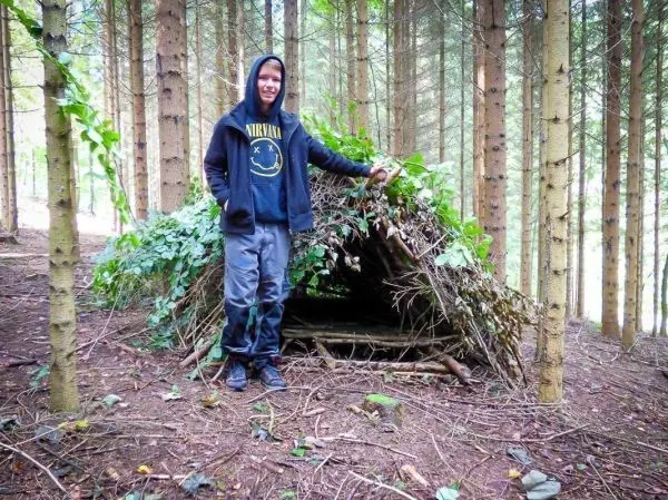 Junge steht neben einer selbstgebauten Tarp-Notunterkunft im Wald