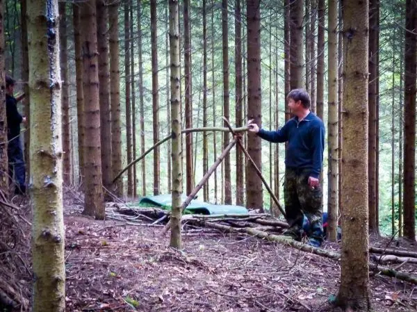 Mann zeigt auf eine selbstgebaute Tarp-Notunterkunft im Wald