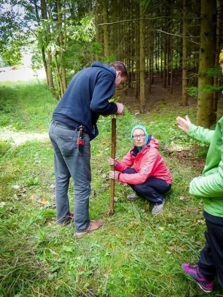 Zwei Personen arbeiten an einem Holzstock im Wald, eine kniet und hält den Stock
