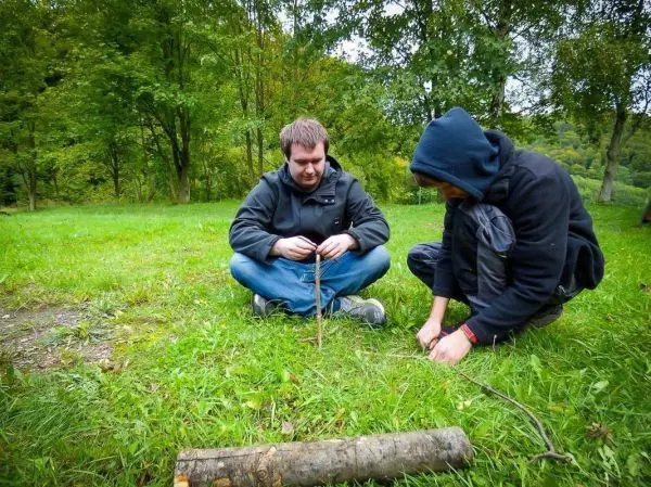 Zwei Personen sitzen im Gras und bearbeiten einen Stock beim Überlebenstraining
