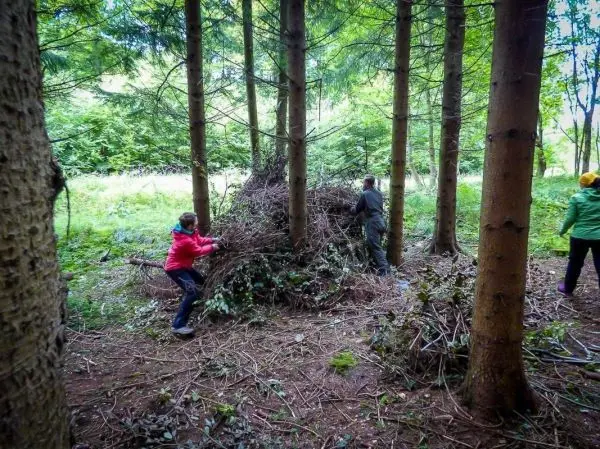 Zwei Personen stapeln Äste und Zweige zu einem Unterschlupf zwischen Bäumen
