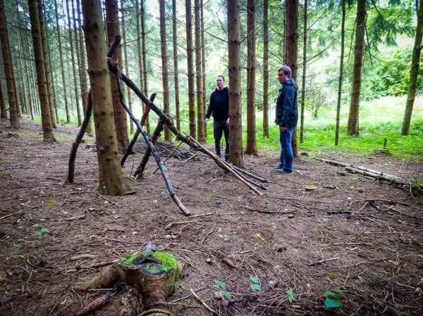 Zwei Personen stehen neben einer selbstgebauten Konstruktion aus Ästen im Wald