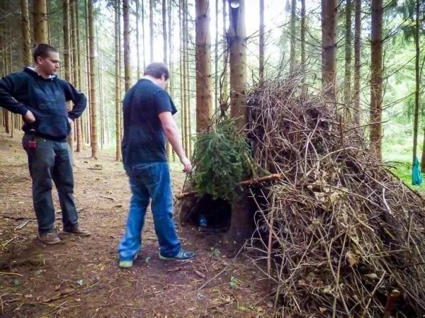 Zwei Personen stehen vor einer selbstgebauten Laubhütte im Wald