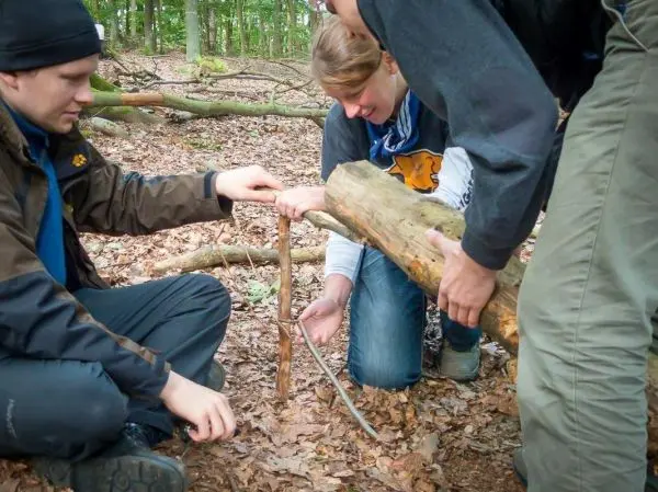 Drei Personen bauen eine Schlagfalle (Figure-Four) aus Holzstücken im Wald