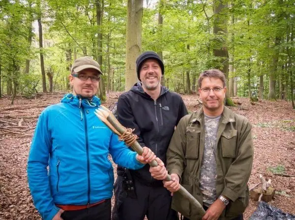 Drei Personen halten gemeinsam einen improvisierten Stock mit einer Spitze in einem Wald