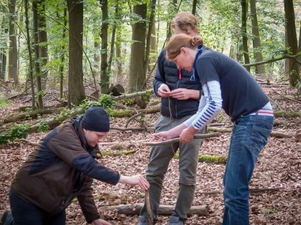 Drei Personen im Wald, eine Person kniet und hält einen Stock