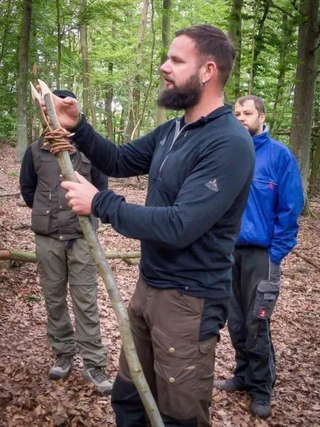 Ein Mann bearbeitet einen Stock mit einem Messer, während zwei Personen im Hintergrund beobachten