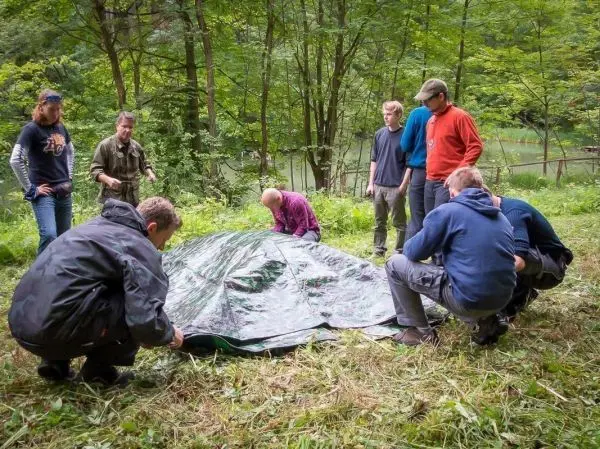 Gruppe baut Tarp-Notunterkunft im Wald auf, mehrere Personen ziehen an den Ecken
