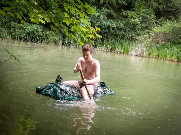 Junge paddelt mit einem Stock auf einer improvisierten Schwimmhilfe im Wasser