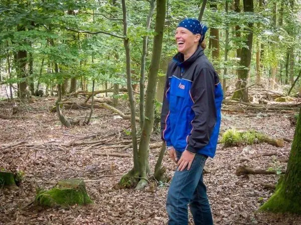 Lächelnde Person steht im Wald zwischen Bäumen und Laub