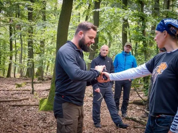 Mann demonstriert eine Technik an einer Frau im Wald, Zuschauer beobachten aufmerksam