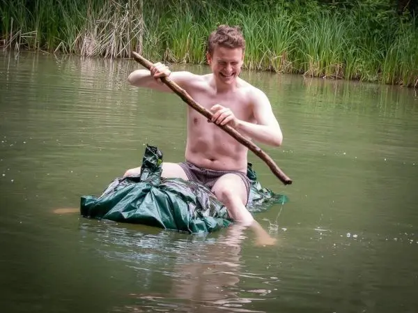 Mann paddelt auf improvisiertem Floß aus Folie mit einem Stock im Wasser