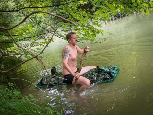 Mann paddelt auf improvisiertem Floß im Wasser mit einem Stock