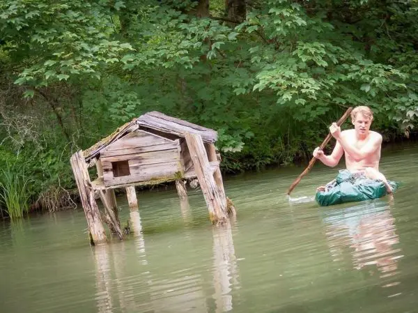 Mann paddelt auf improvisiertem Floß in einem Gewässer neben einer hölzernen Struktur
