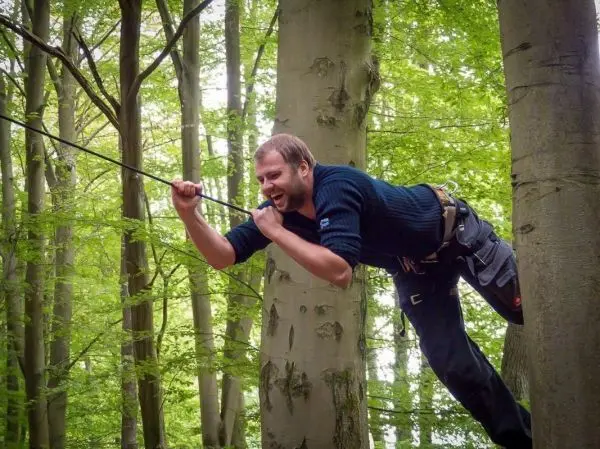 Mann überquert mit Seil eine Baumkronenverbindung zwischen zwei Bäumen