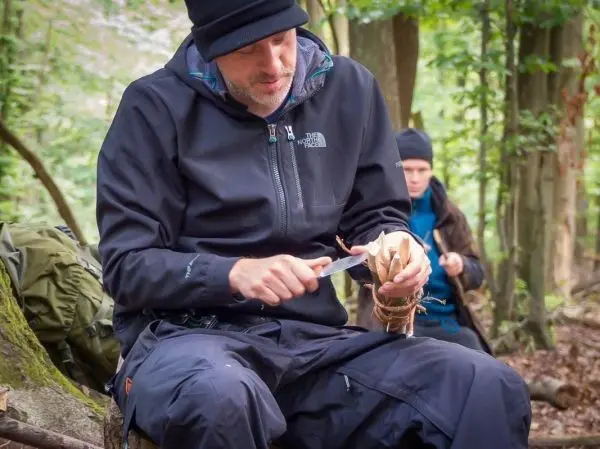 Messer schnitzt Holzstück während jemand im Hintergrund sitzt