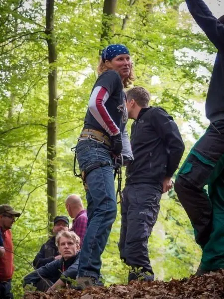 Person mit Kletterausrüstung steht im Wald, umgeben von weiteren Personen