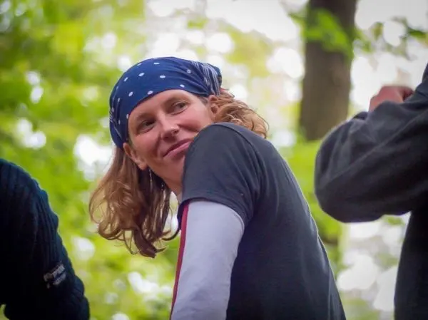 Person mit Kopftuch und T-Shirt lächelt in die Kamera, umgeben von Bäumen