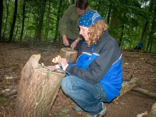 Zunder wird mit einem Feuerstahl entzündet, während eine zweite Person Holz bearbeitet